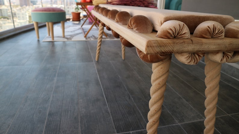 May include: A wooden bench with a beige cushion and rope legs. The cushion is decorated with brown velvet buttons.