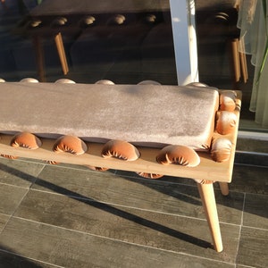 May include: A wooden bench with a light brown cushion and brown tufted detailing. The bench has four legs and is made of natural wood.