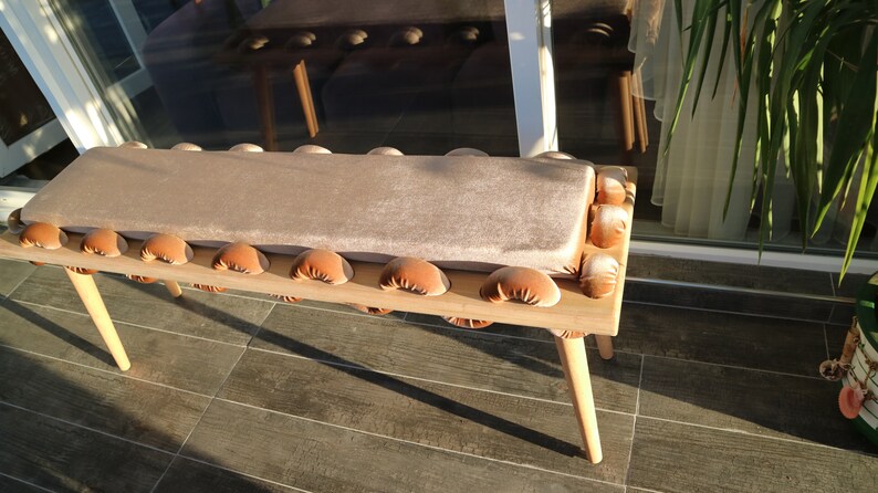May include: A wooden bench with a light brown cushion and brown tufted detailing. The bench has four legs and is made of natural wood.