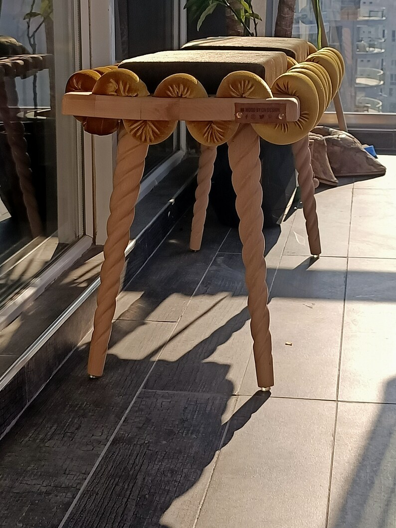 May include: A wooden bench with a rope-like design on the legs. The bench has a padded seat with a row of yellow velvet buttons. The bench is labeled "MADE BY CO. DESIGN".