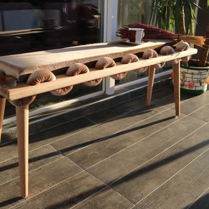 May include: A wooden bench with a padded seat and rounded cushions. The bench has a natural wood finish and is decorated with a light brown fabric.