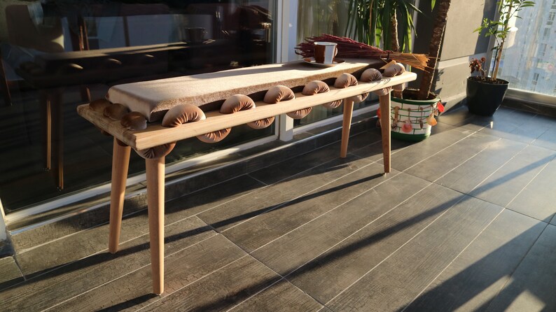 May include: A wooden bench with a padded seat and rounded cushions. The bench has a natural wood finish and is decorated with a light brown fabric.