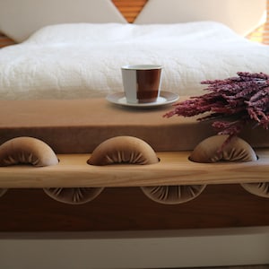 May include: A wooden bench with a brown velvet cushion and rope legs. The bench has a series of rounded, cushioned supports along the top. A cup of coffee and a bouquet of dried flowers are on the bench.
