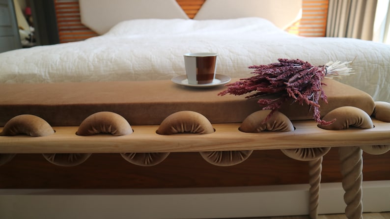 May include: A wooden bench with a brown velvet cushion and rope legs. The bench has a series of rounded, cushioned supports along the top. A cup of coffee and a bouquet of dried flowers are on the bench.