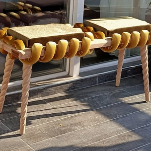 May include: A wooden bench with two cushioned seats. The seats are covered in a mustard yellow fabric with a rope-like design. The bench has four twisted legs.