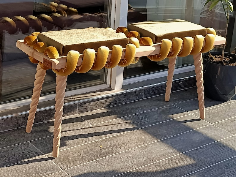 May include: A wooden bench with two cushioned seats. The seats are covered in a mustard yellow fabric with a rope-like design. The bench has four twisted legs.