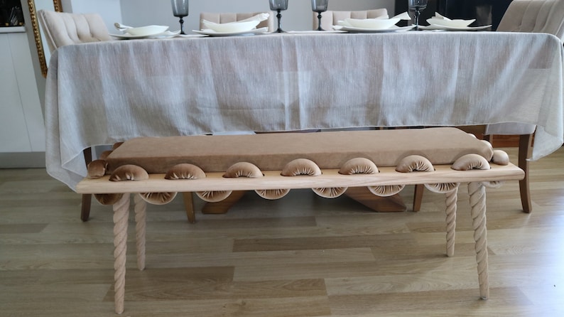 May include: A wooden bench with a brown velvet cushion and rope legs. The bench has a unique design with rounded edges and a series of small, rounded cushions along the front.
