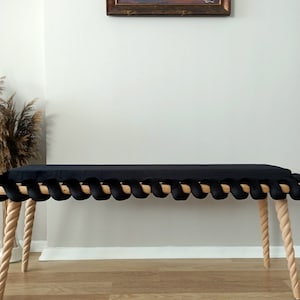 Può includere: Una panca in velluto nero con un design a corda sul bordo anteriore. La panca ha quattro gambe in legno intrecciate e una finitura in legno naturale.