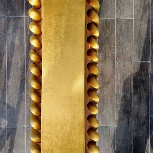 K&ouml;nnte beinhalten: Ein rechteckige, senfgelbe Samtbank mit Holzrahmen. Die R&auml;nder sind mit einer Reihe von goldenen, abgerundeten, dekorativen Elementen verziert. Die Bank steht vor einem Hintergrund aus grauem Fliesenboden. Ma&szlig;e: ca. 120 cm lang und 40 cm breit.
