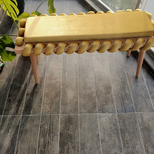 K&ouml;nnte beinhalten: Eine goldfarbene Bank mit einem rechteckigen Kissen und Zierleisten. Die Bank hat helle Holzbeine und einen hellen Holzrahmen. Die Zierleiste besteht aus einem gedrehten, seilartigen Material. Die Bank steht auf einem Holzboden.