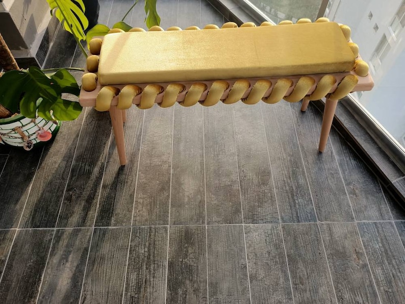 K&ouml;nnte beinhalten: Eine goldfarbene Bank mit einem rechteckigen Kissen und Zierleisten. Die Bank hat helle Holzbeine und einen hellen Holzrahmen. Die Zierleiste besteht aus einem gedrehten, seilartigen Material. Die Bank steht auf einem Holzboden.