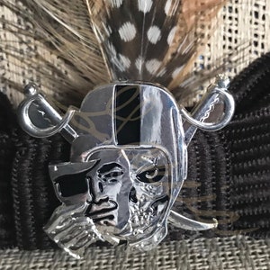 May include: A silver skull and crossbones pin with a black stripe across the skull. The pin is detailed with a pirate's face and a skull.