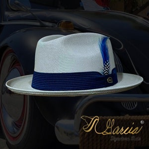 May include: A white straw fedora with a blue band and a blue feather accent. The hat is sitting on a vintage car.
