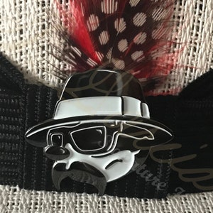 May include: A black and white enamel pin of a cartoon character wearing a fedora and sunglasses. The character has a moustache and a goatee. The pin is attached to a black and white striped fabric.