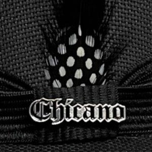 Könnte beinhalten: Schwarzer Hut mit einem silbernen "Chicano"-Logo und einem schwarzen und weißen Tupfen-Federbesatz.
