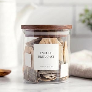 May include: A clear glass jar with a wooden lid, filled with tea bags. The jar has a white label that reads "ENGLISH BREAKFAST" and "PREMIUM QUALITY". A wooden spoon and a white teapot are in the background.