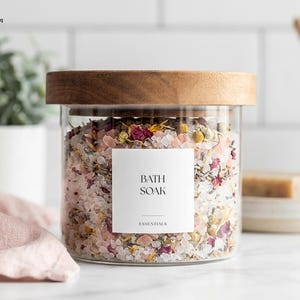 Może przedstawiać: Przezroczysty szklany słoik z drewnianą pokrywką wypełniony solą do kąpieli. Słoik ma białą etykietę z napisem "BATH SOAK" i "ESSENTIALS". Sól do kąpieli to mieszanka różowych, białych i żółtych suszonych kwiatów i soli.