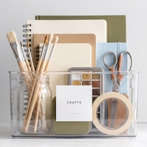 May include: A clear plastic bin filled with craft supplies. The bin contains paintbrushes, notebooks, a jar, scissors, a palette of paints, and a roll of tape. A white label reads "CRAFTS".