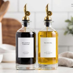 Op de afbeelding: Twee heldere glazen flessen met gouden tuiten, &eacute;&eacute;n gevuld met donkere balsamico-azijn en de andere met olijfolie. Elke fles heeft een wit etiket met zwarte tekst. Het ontwerp is signature, en de labelgrootte is 6,35 cm x 7,62 cm.
