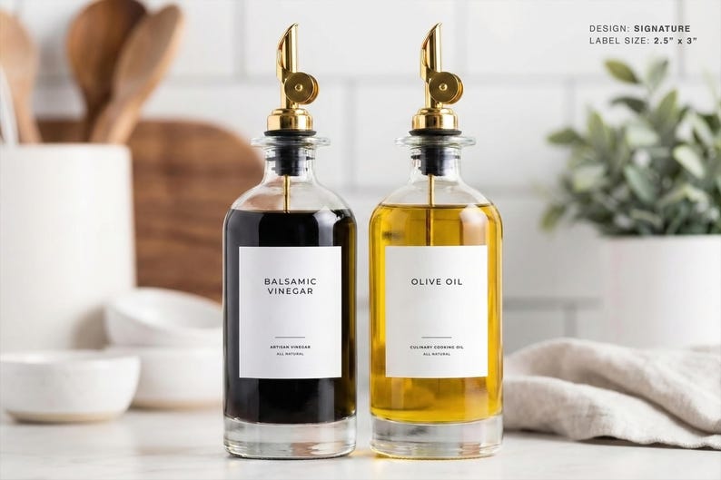 Op de afbeelding: Twee heldere glazen flessen met gouden tuiten, &eacute;&eacute;n gevuld met donkere balsamico-azijn en de andere met olijfolie. Elke fles heeft een wit etiket met zwarte tekst. Het ontwerp is signature, en de labelgrootte is 6,35 cm x 7,62 cm.