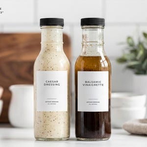 Op de afbeelding: Twee glazen flessen saladedressing met zwarte doppen. E&eacute;n bevat Caesar dressing, de andere balsamico vinaigrette. Elke fles heeft een wit etiket met zwarte tekst. Het etiket is 6,35 cm x 7,62 cm groot.