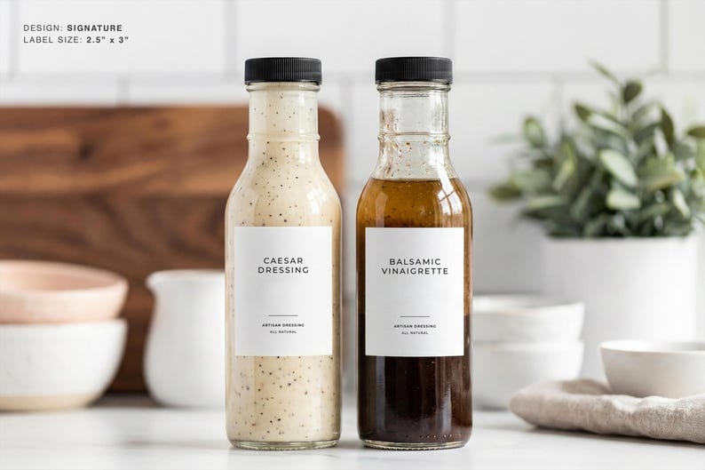 Op de afbeelding: Twee glazen flessen saladedressing met zwarte doppen. E&eacute;n bevat Caesar dressing, de andere balsamico vinaigrette. Elke fles heeft een wit etiket met zwarte tekst. Het etiket is 6,35 cm x 7,62 cm groot.