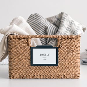 May include: A rectangular woven seagrass basket filled with folded fabric towels in solid cream, grey striped, and white plaid patterns. A black rectangular clip-on label holder is attached to the front, displaying the white text TOWELS KITCHEN.
