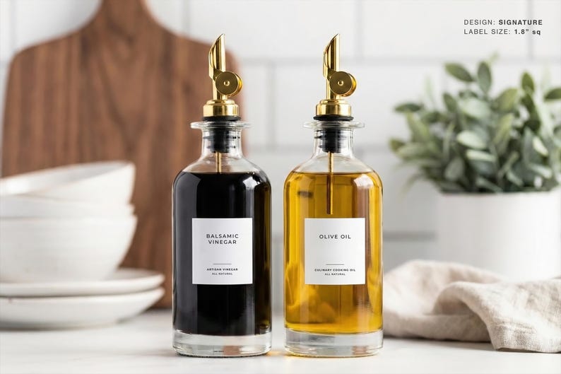 Op de afbeelding: Twee glazen flessen met goudkleurige schenktuiten, &eacute;&eacute;n gevuld met donkere balsamicoazijn en de andere met olijfolie. Elke fles heeft een wit etiket met zwarte tekst. De balsamicoazijnfles is gelabeld met "Balsamicoazijn", en de olijfoliefles met "Olijfolie".