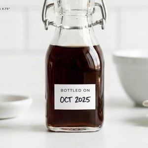 May include: A small glass bottle filled with a dark liquid, sealed with a metal clasp. A white label on the front reads "BOTTLED ON OCT 2025". The bottle is on a white surface, with a white bowl and folded cloth in the background.