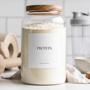 Puede incluir: Un gran tarro de vidrio con tapa de madera, lleno de proteína en polvo. Una etiqueta blanca en el tarro dice "PROTEÍNA" y "SUPLEMENTO". Otros artículos de fitness están en el fondo.