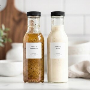 Op de afbeelding: Twee glazen flessen met zwarte doppen, een gevuld met Italiaanse dressing en de andere met knoflook aioli. Elke fles heeft een wit etiket met zwarte tekst. De Italiaanse dressing is bruinachtig groen en de knoflook aioli is wit.