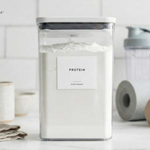 May include: A clear, rectangular container filled with white powder, labeled "PROTEIN SUPPLEMENT." The container is part of a minimalist design, with a label size of 3" x 2.5". Other items in the background include a mug, a yoga mat, and dumbbells.