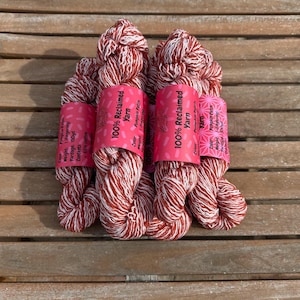 May include: Four skeins of red and white reclaimed yarn, each wrapped with a pink label. The labels read "100% Reclaimed Yarn" and include details about the yarn's weight and content. The yarn is displayed on a wooden surface.