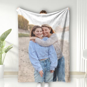 May include: A photo of two people hugging on a beach. The person in the front is wearing a blue shirt and jeans. The person in the back is wearing a white shirt and jeans.