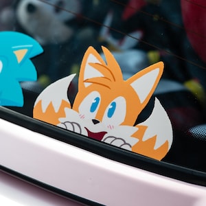 May include: A cartoon sticker of Tails, a yellow fox character from the Sonic the Hedgehog video game series, peeking out of a car window. Tails has a big smile and blue eyes.