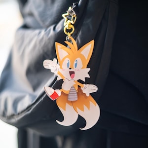 May include: A yellow and white Tails the Fox keychain with a gold-colored star-shaped clasp. The cartoon character is smiling with its mouth open and one leg raised. The keychain is attached to a black backpack.