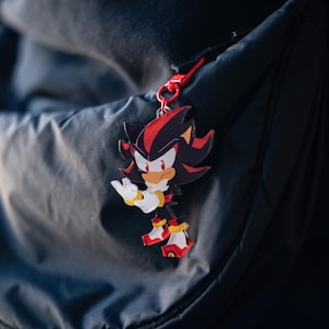 May include: A colorful Shadow the Hedgehog keychain. The keychain features the character in a dynamic pose, with a red and black color scheme. The keychain is attached to a red clasp.