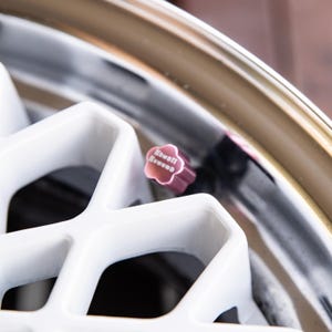 Peut inclure: Gros plan sur une roue de voiture avec un design à rayons blancs et un bouchon de valve rose. Le bouchon porte l'inscription "Kawaii Rununo" en blanc. La jante de la roue est dorée.