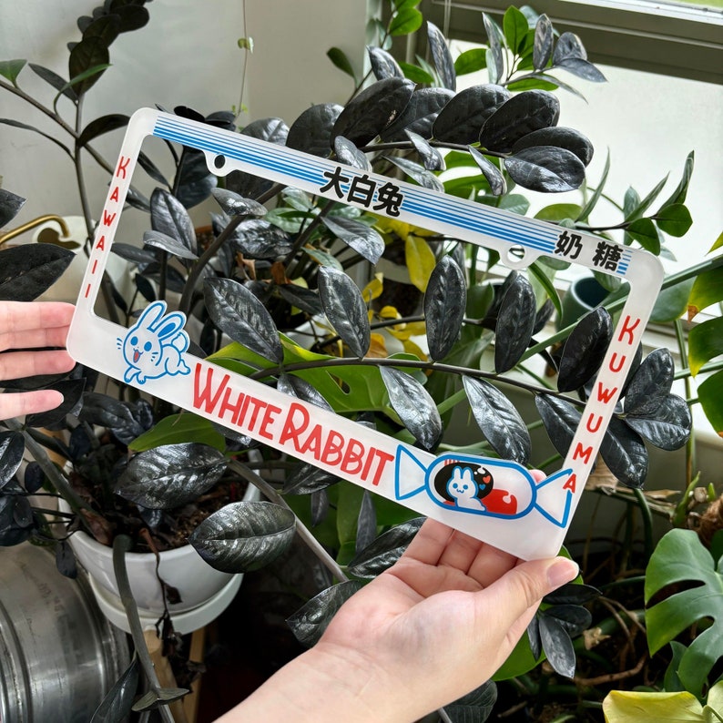 以下が含まれることがあります： 白いナンバープレートフレーム。漫画のウサギと赤い「White Rabbit」の文字が特徴です。フレームには日本語のテキストとピンク色の「Kawaii」という単語も含まれています。