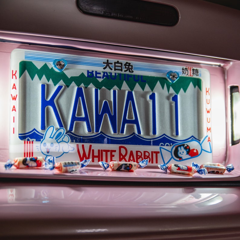 以下が含まれることがあります： 青色の「KAWAII」という文字と数字の「11」が入った装飾的なナンバープレート。プレートは白と緑のデザインで、「BEAUTIFUL」と「WHITE RABBIT」という文字が入っています。プレートの前にはいくつかのキャンディーが散らばっています。