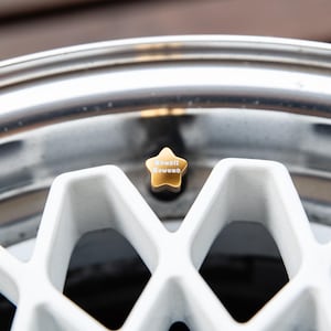 May include: Close-up of a car wheel with a white geometric design and a shiny silver rim. A gold star-shaped valve stem cap is centered, bearing the text "Kawaii Kuwuno".