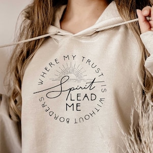 Puede incluir: Una sudadera con capucha beige con un estampado gráfico negro que dice "Where my trust is without borders, Spirit lead me". El gráfico también incluye un sol estilizado con rayos.