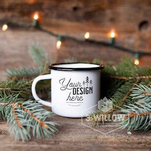 May include: White enamel mug with black rim and handle, featuring the text "Your Design Here" and a tree graphic. The mug is surrounded by pine branches and fairy lights, set against a rustic wooden background.