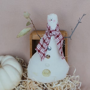 May include: A white fabric snowman with a red and white checked scarf and brown twig arms. The snowman is sitting in a wooden box filled with shredded paper and next to a white pumpkin.