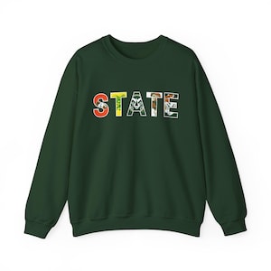May include: A dark green sweatshirt with the word "STATE" printed across the chest. Each letter is filled with a different colorful design, including a tiger, a leaf, and other patterns. The sweatshirt has long sleeves and a crew neck.