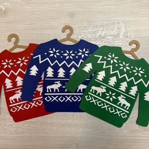 May include: Three paper Christmas sweater ornaments in red, blue, and green with white snowflake and reindeer designs. The sweaters are cut out and have a hanger at the top.