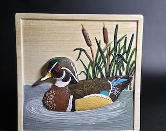 May include: A square wooden plaque featuring a relief carving of a wood duck swimming in gray water. The duck has a green, white, and brown head with a red eye, a brown body, and yellow and blue wing details. Green cattails rise in the background.