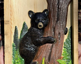 May include: A rectangular wooden wall plaque featuring a three-dimensional relief carving of a black bear cub climbing a brown tree trunk. The scene includes painted green pine trees, ferns, and a small white flower against a light wood background.