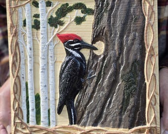 May include: A square wooden plaque featuring a relief carving of a black woodpecker with a red crest perched on a dark brown tree trunk. The scene includes white birch trees with green foliage in the background and a carved vine border frame.