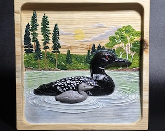 May include: A square wooden plaque featuring a carved and painted scene of a black loon with a small gray chick swimming in water. The background displays a forest of green pine trees, a deciduous tree, a yellow sun, and light clouds.
